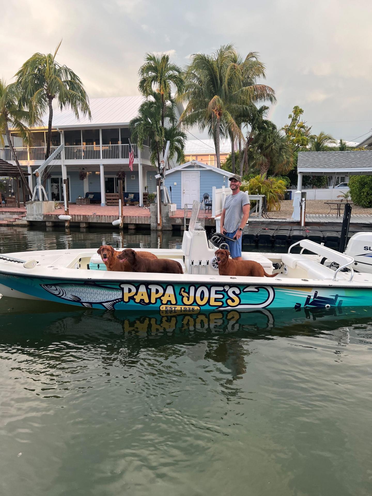 A person and two dogs on a boat named "PAPA JOE'S" docked by palm trees.