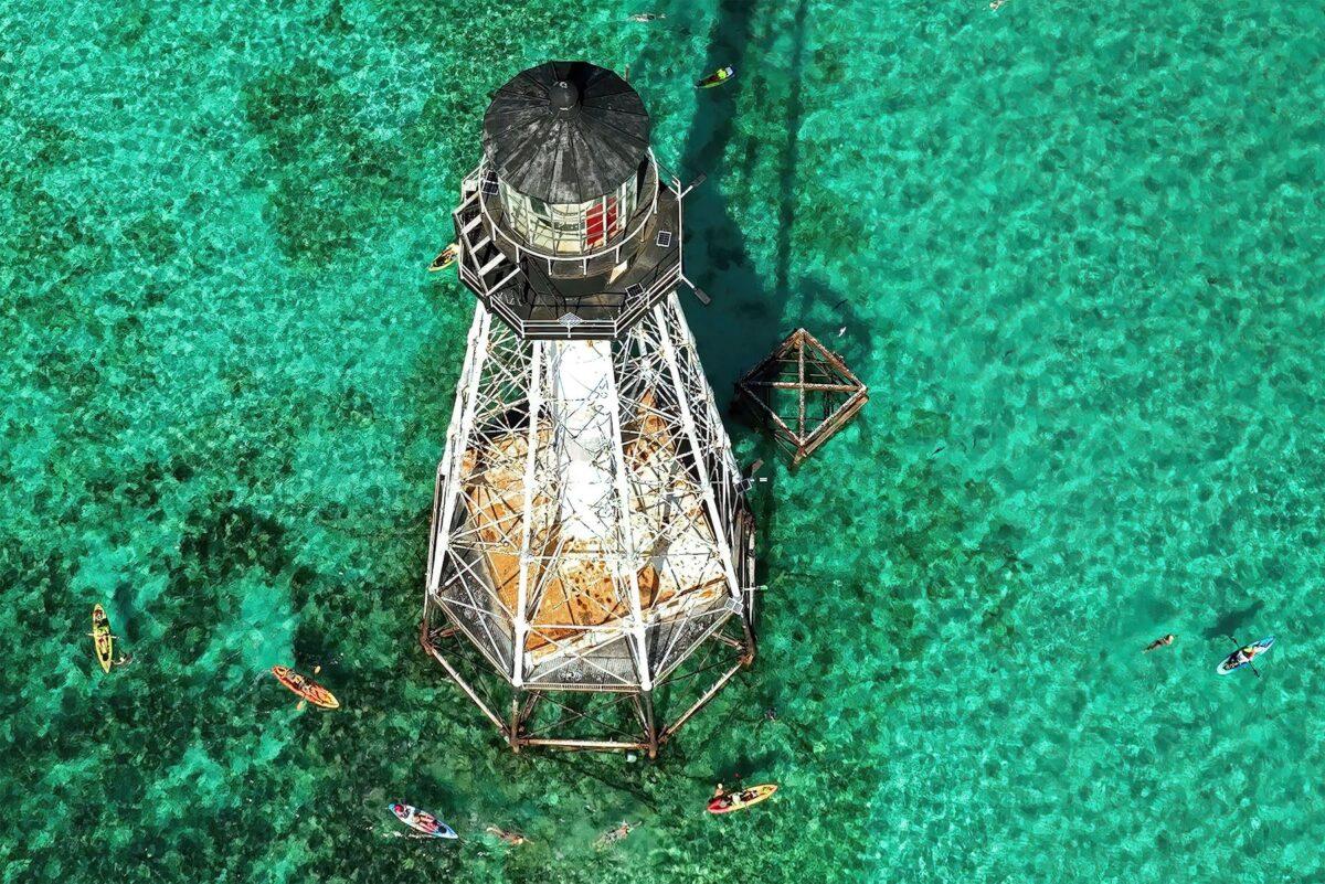 Lighthouse over clear turquoise water, surrounded by small boats.