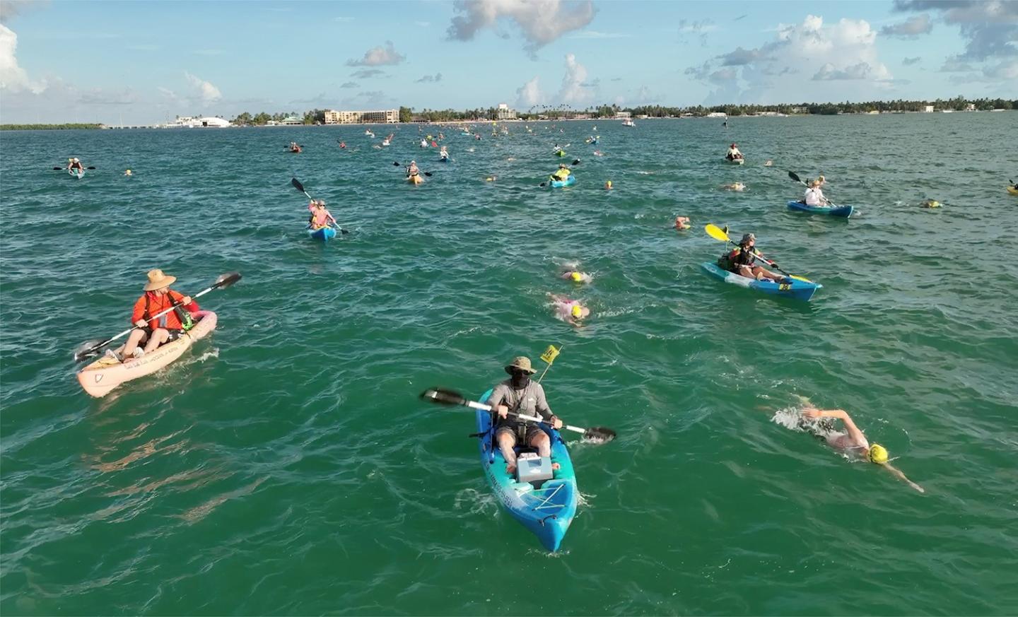 Kayakers and swimmers on a wide expanse of open water under a bright blue sky.