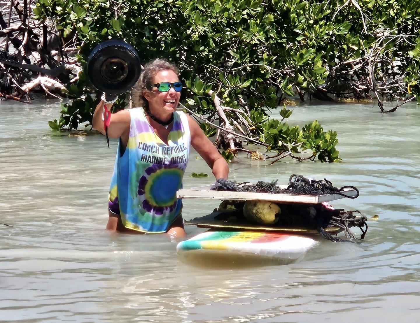 Woman in tie-dye shirt wading in water, holding a pan and surfboard.