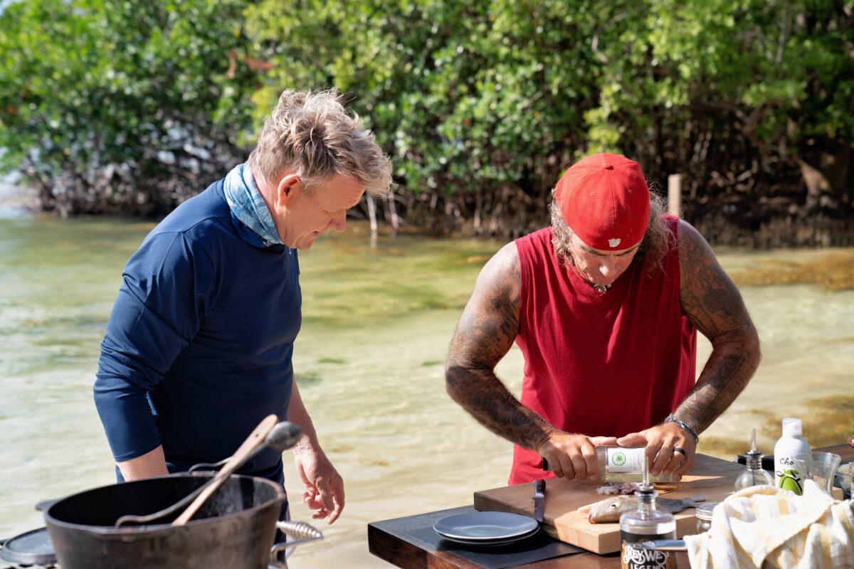 Two men cooking outdoors by the water, surrounded by greenery.