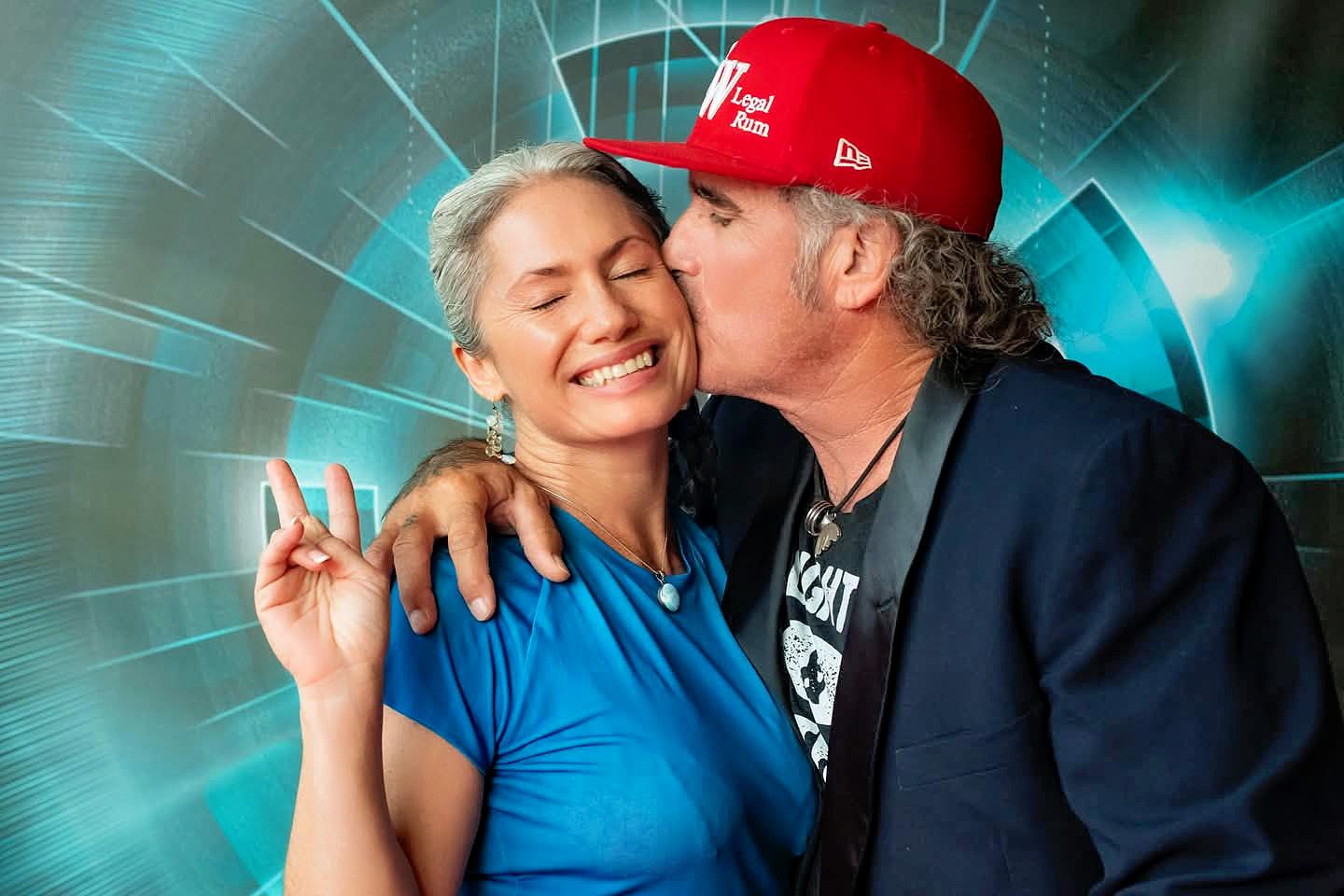 Man kisses smiling woman posing with peace sign; vibrant backdrop.