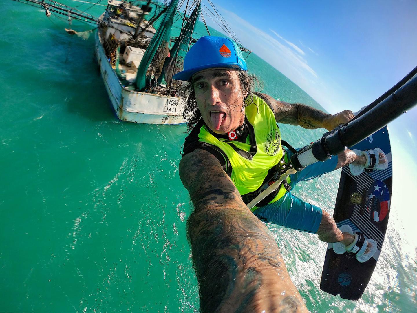Kiteboarder in blue helmet over turquoise ocean, tongue out, fishing boat in background.