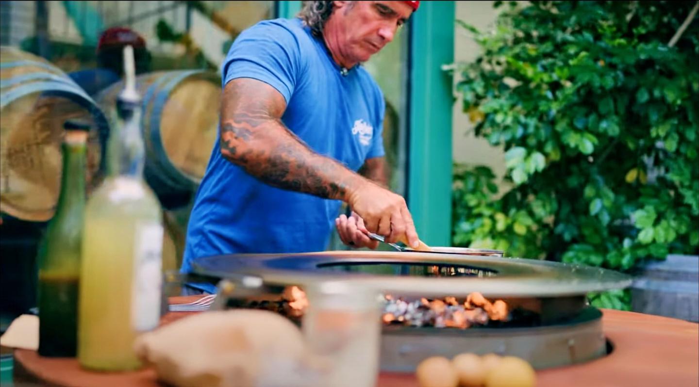 Man grilling food outdoors with barrels and greenery in the background.