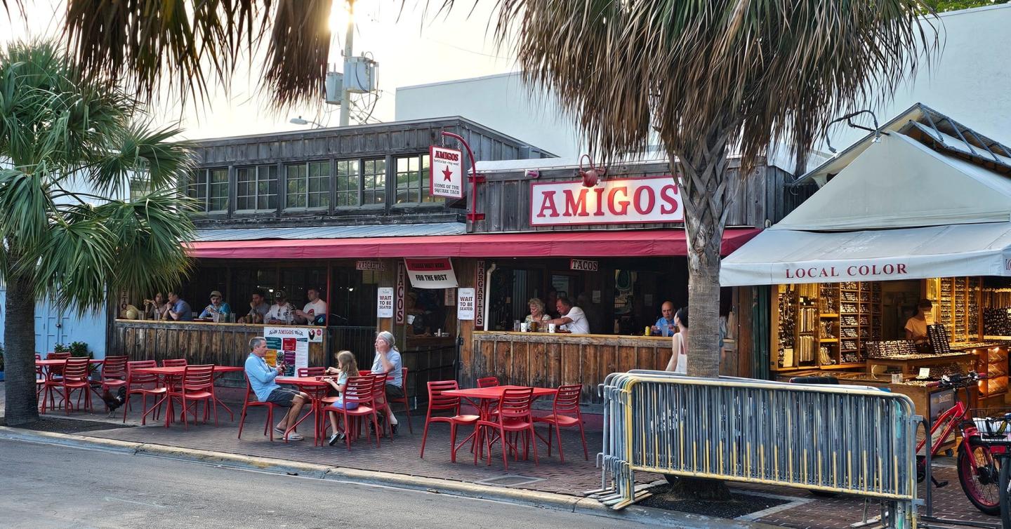 Outdoor restaurant with red chairs, patrons dining, palm trees, and colorful awning.