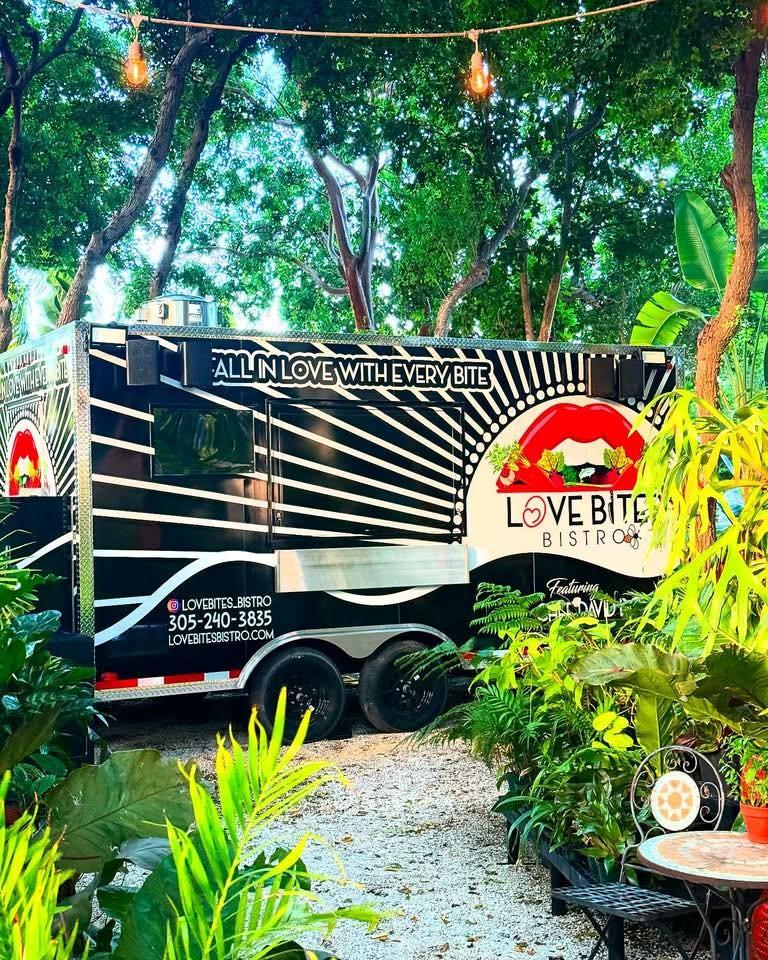 Food truck among lush green plants and trees, colorful and inviting setting.