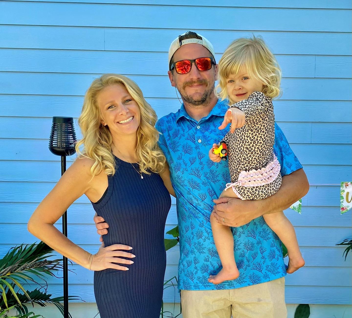 Family smiling in front of a blue wall; man holding child.