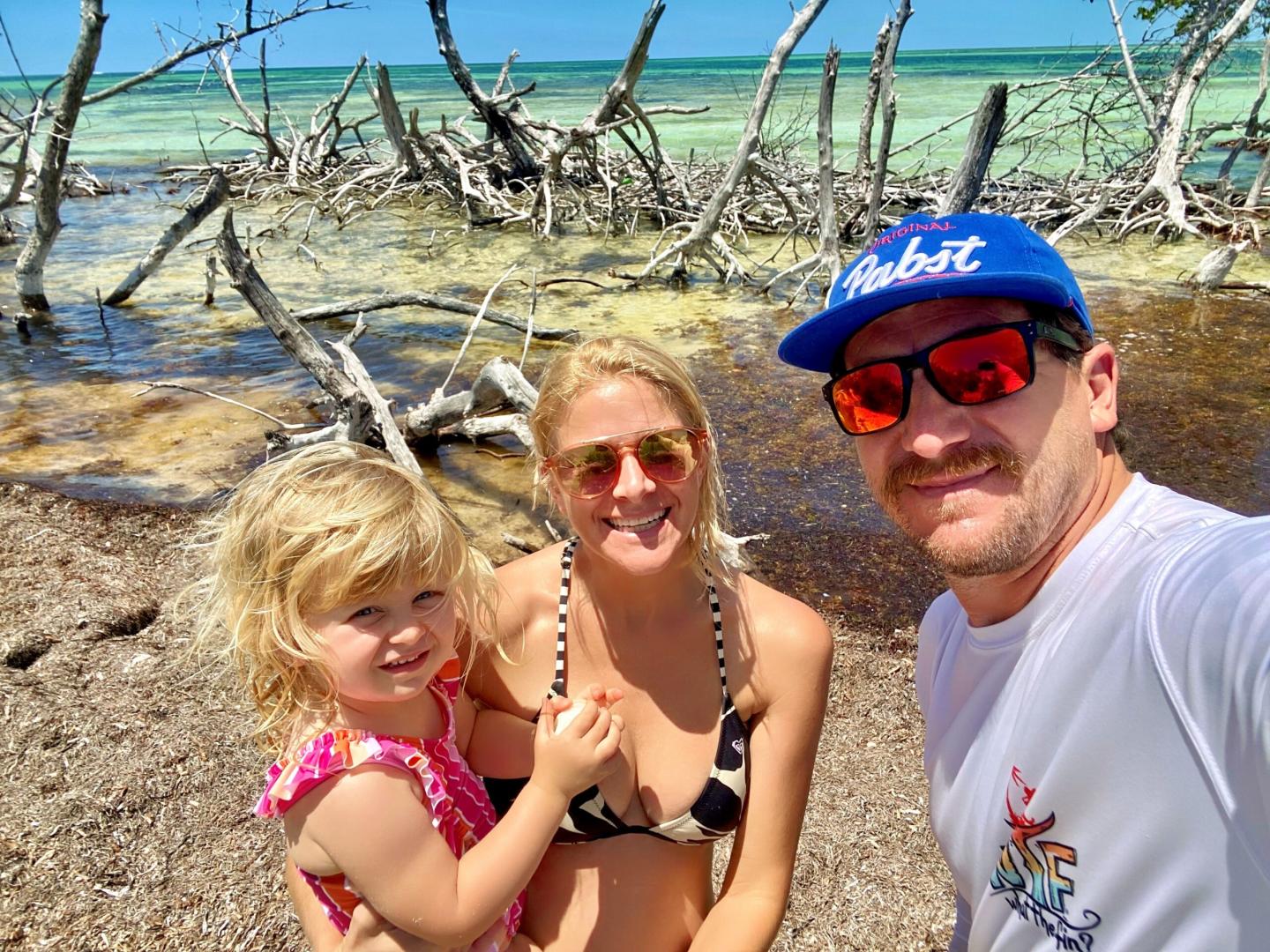 Family smiling at a beach with driftwood and turquoise water.