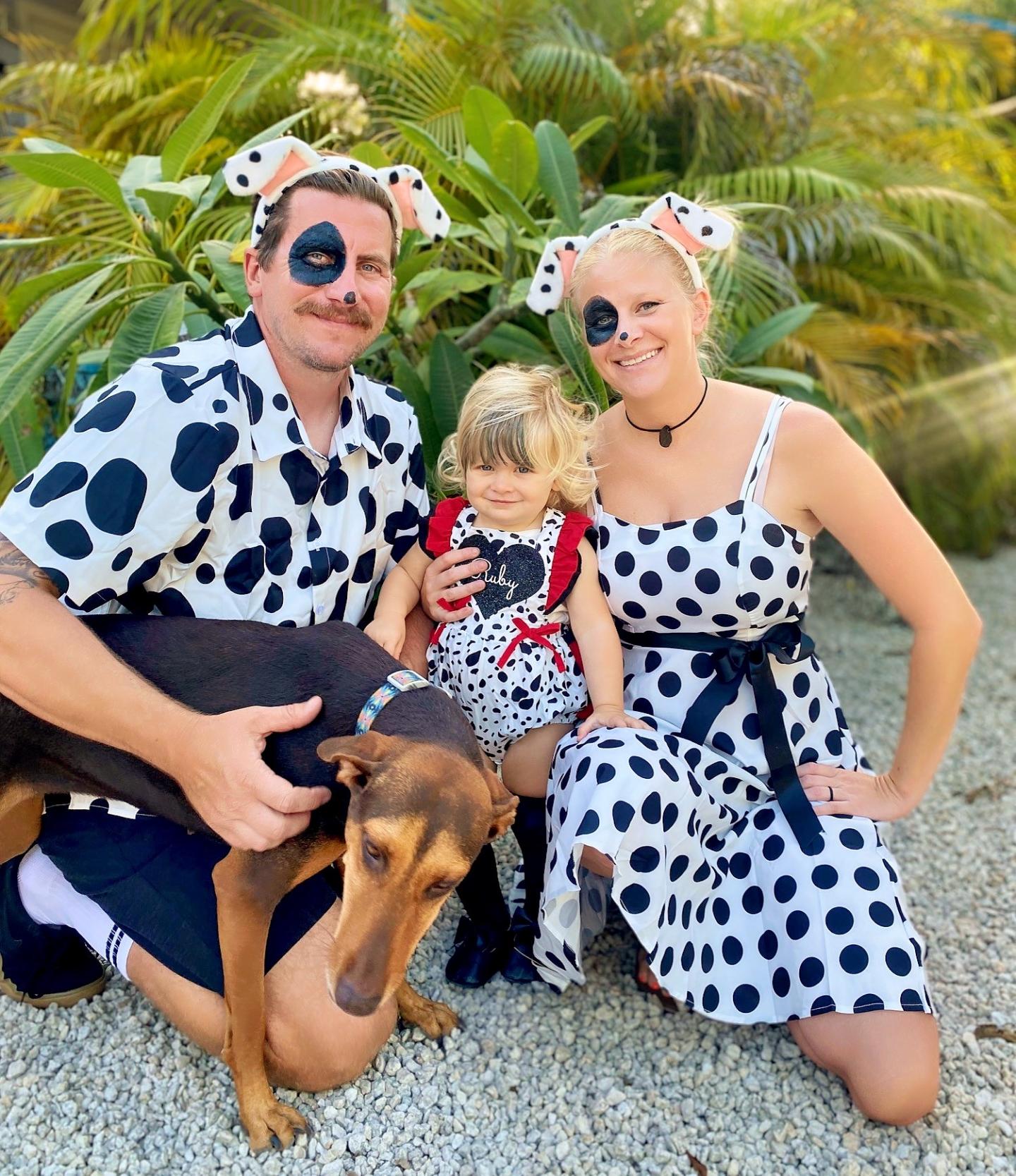 Family in matching dalmatian costumes with a dog, posing outdoors among green plants.