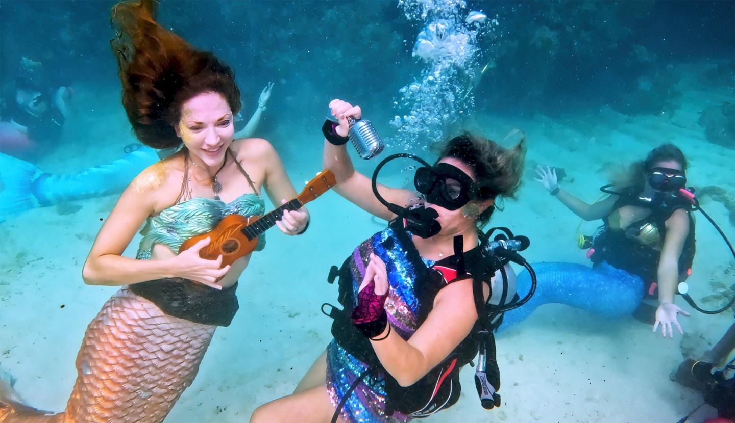 Mermaid and divers playing music underwater, surrounded by clear blue water.