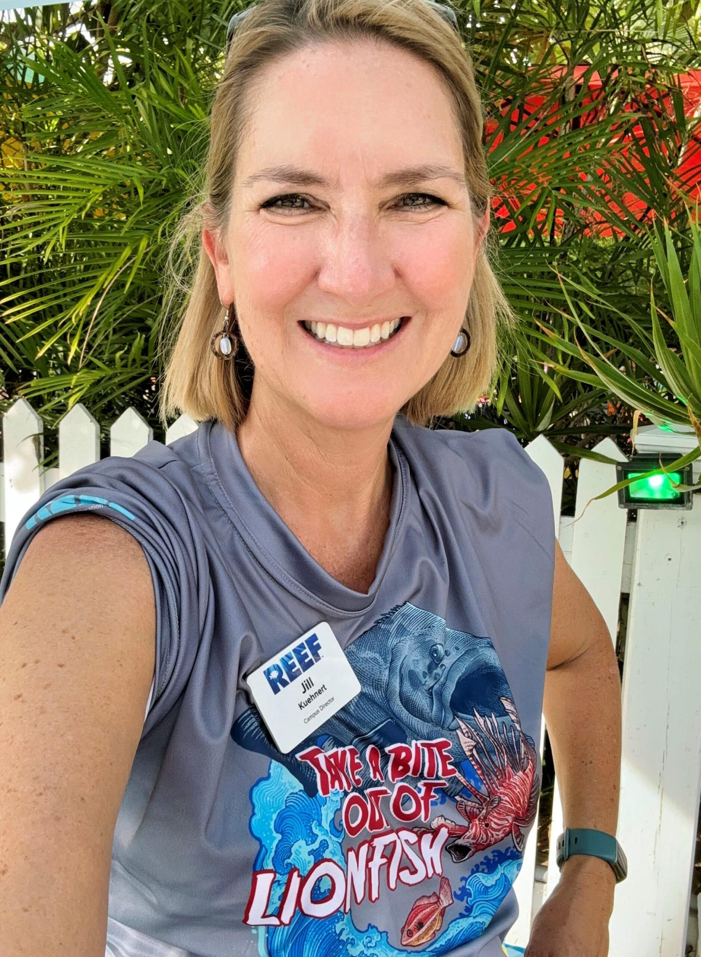 Smiling woman in a gray sleeveless shirt, tropical background.