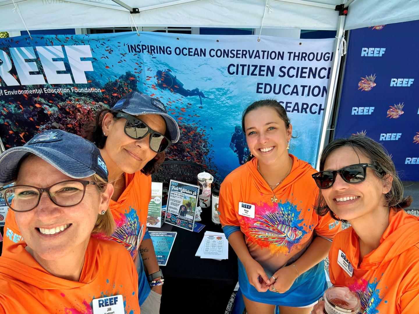 Four women smiling at an outdoor event booth, wearing colorful shirts and sunglasses.