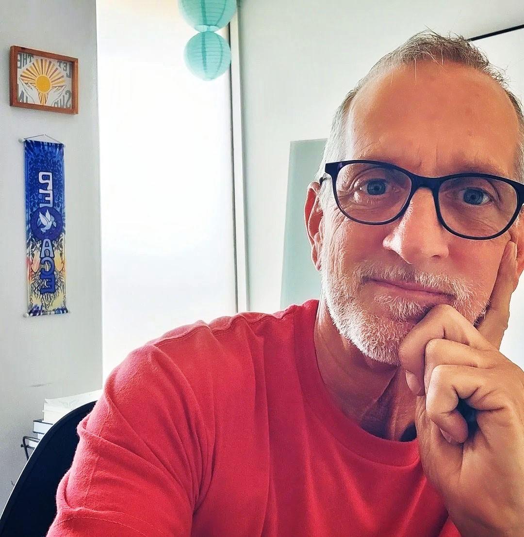 Man wearing glasses and a red shirt, sitting and looking into the camera thoughtfully.