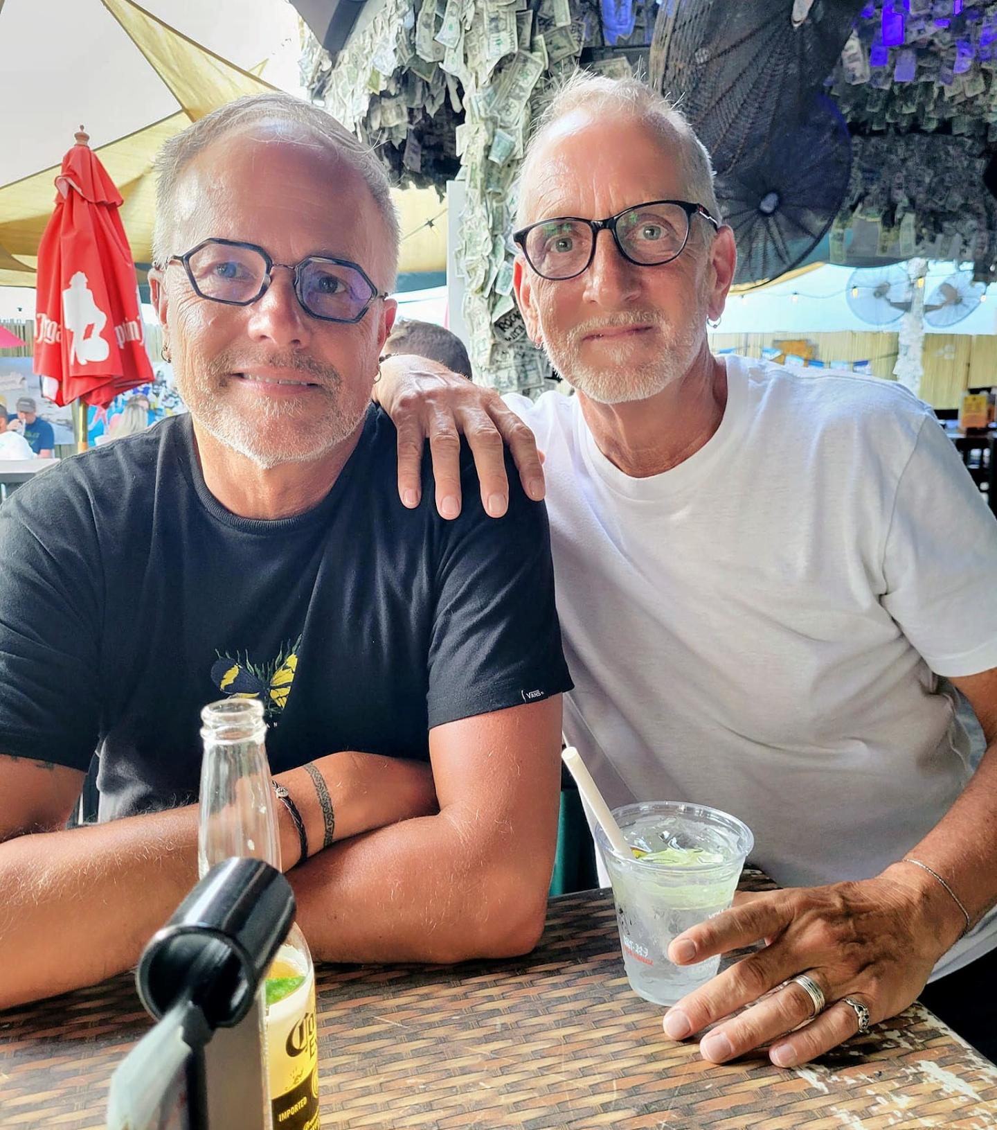 Two smiling men with glasses at an outdoor restaurant.