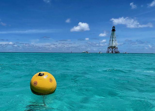 Yellow buoy floating in clear turquoise sea with distant lighthouse under blue sky.