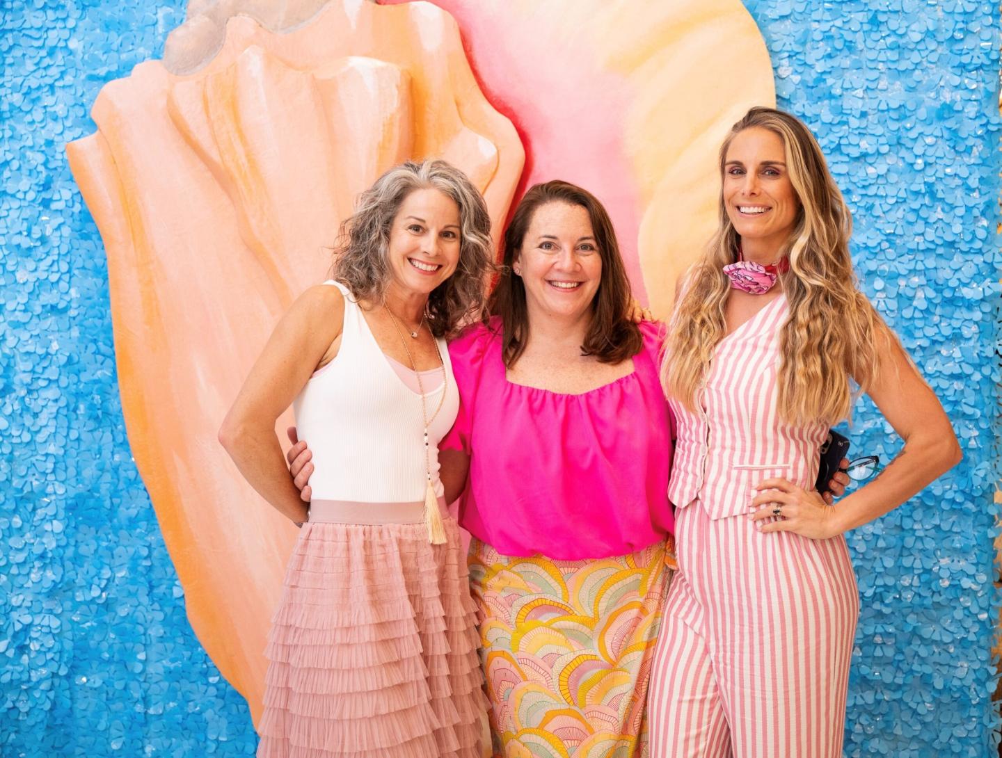 Three women smiling in front of a colorful abstract background.
