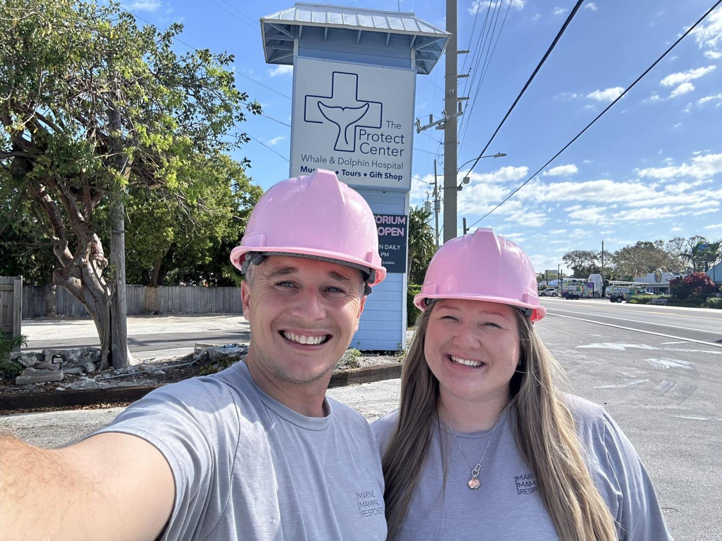 Two people smiling in pink hard hats near a sign, under a clear blue sky.