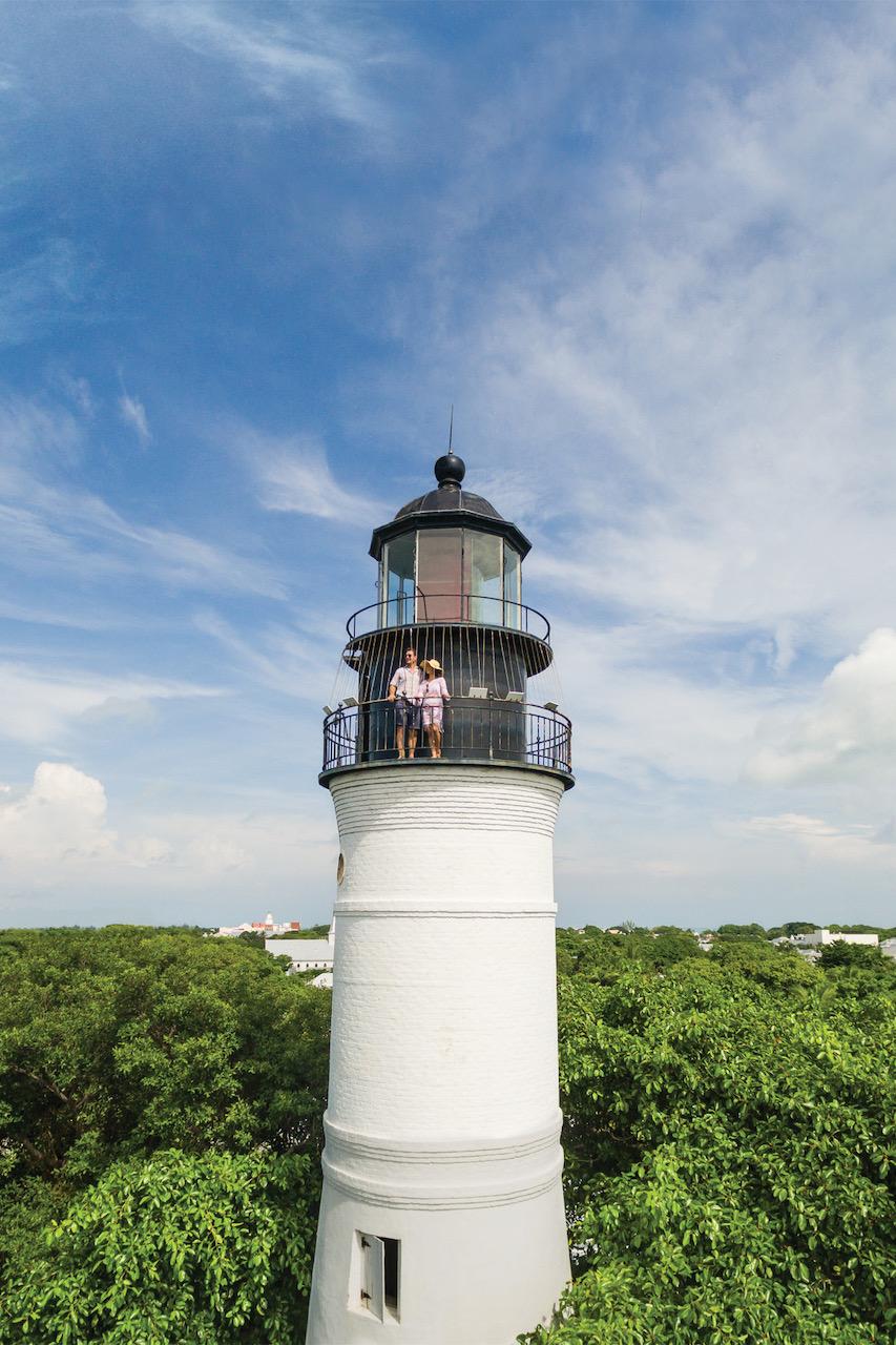 White lighthouse against a blue sky, surrounded by lush green trees.