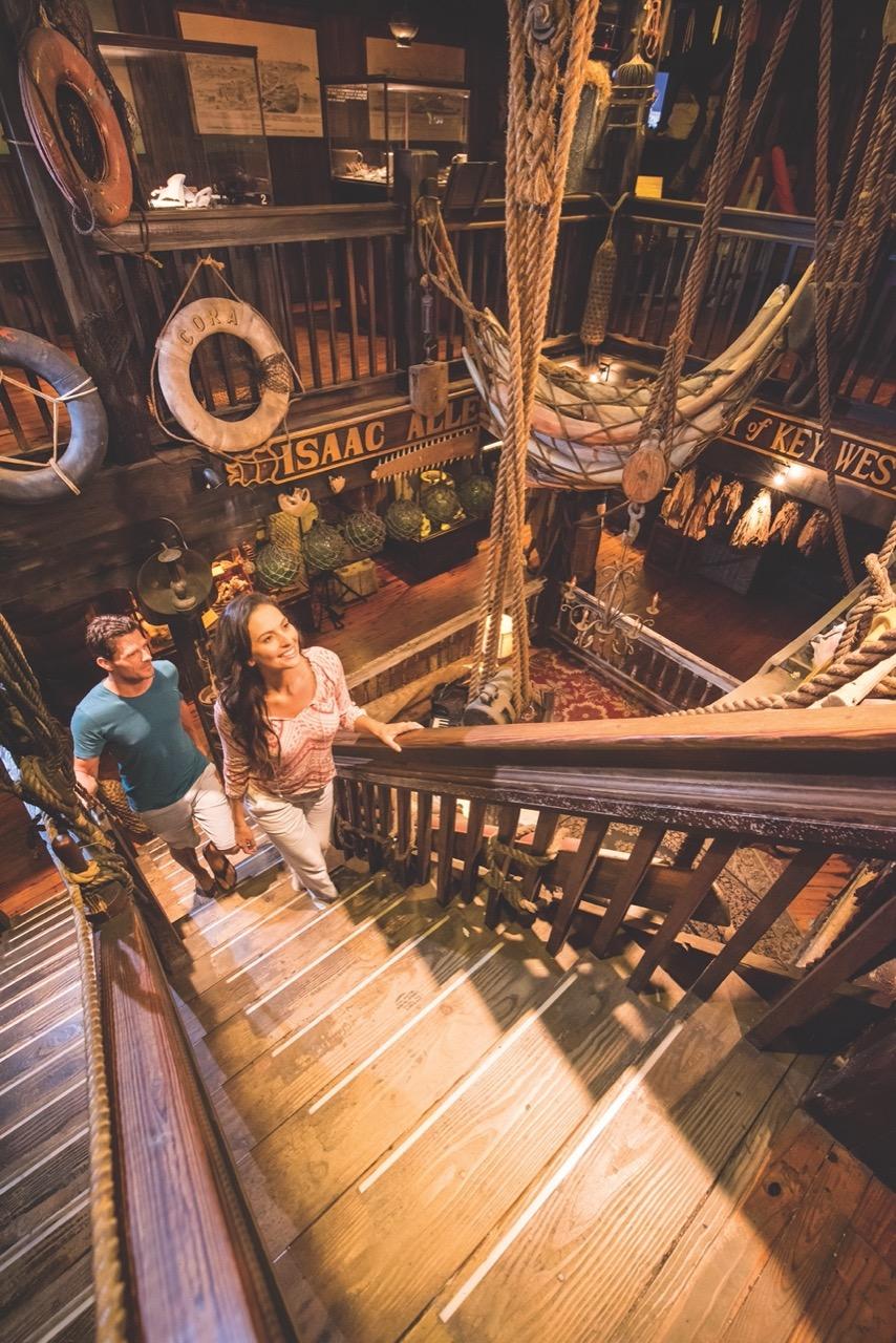 People ascending wooden stairs in a maritime-themed room with lifebuoys and ropes.