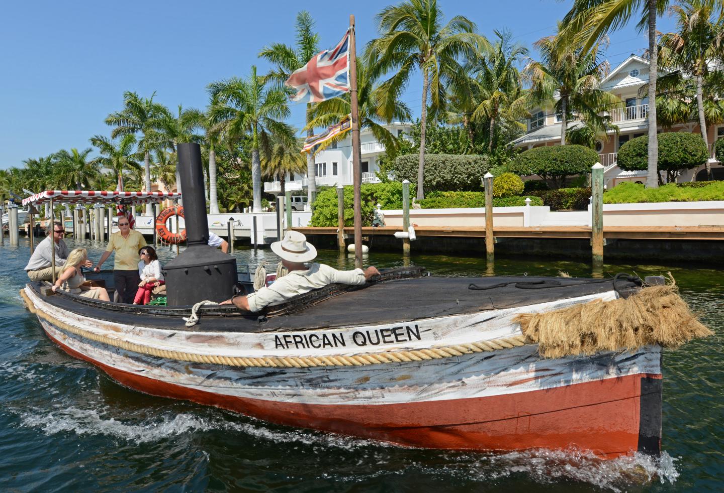 Small boat with passengers cruising near palm-lined canal homes.