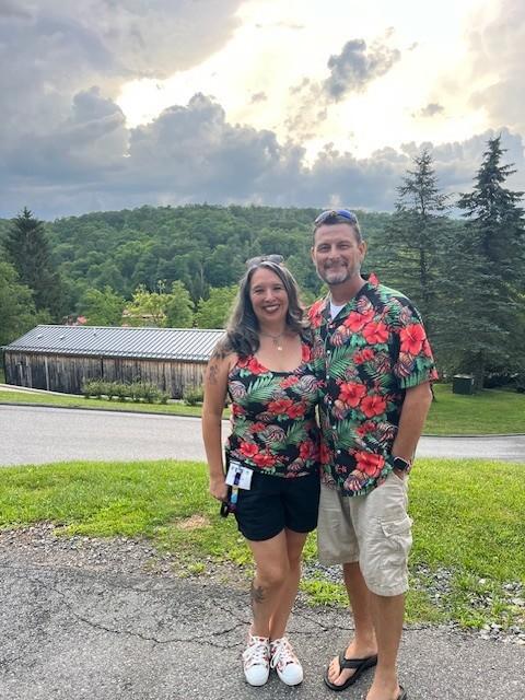 Couple in matching floral shirts, smiling, with trees and cloudy sky in the background.