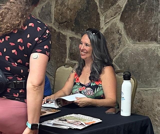 Woman at a table signing books, smiling at someone standing.