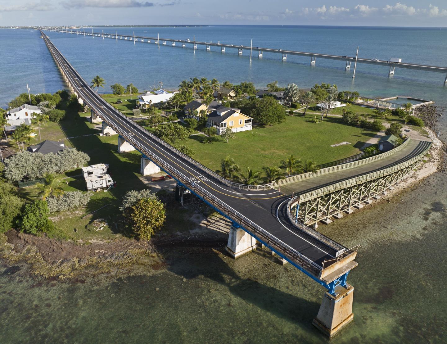 Bridge extends over blue water toward small island with houses and greenery.