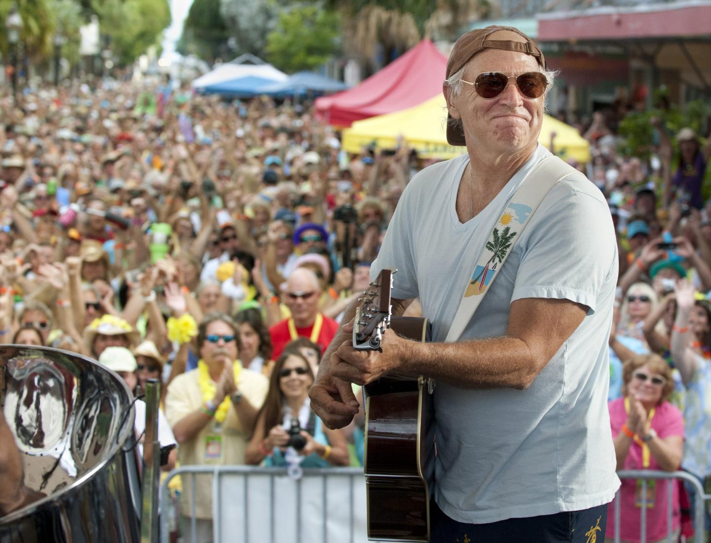 Musician playing guitar on stage before a large outdoor crowd.