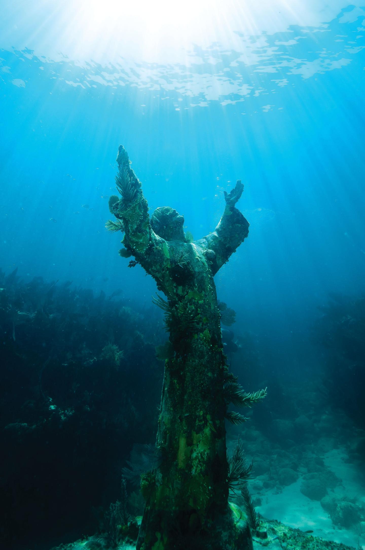 Underwater statue of Christ with arms raised, bathed in blue light.