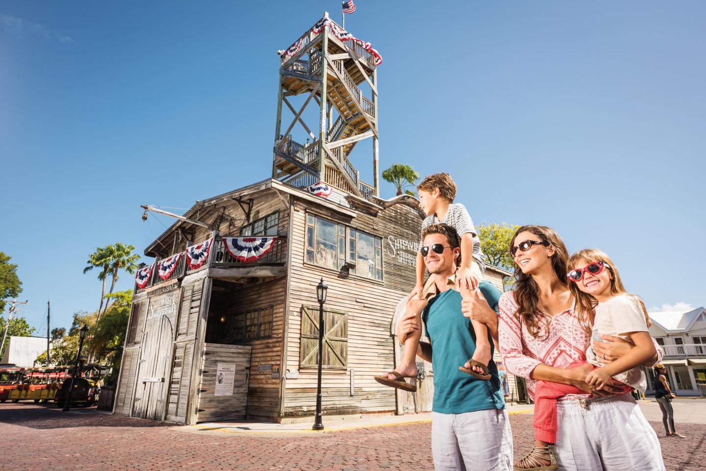 Family in sunglasses poses by rustic wooden building with tower, clear blue sky above.