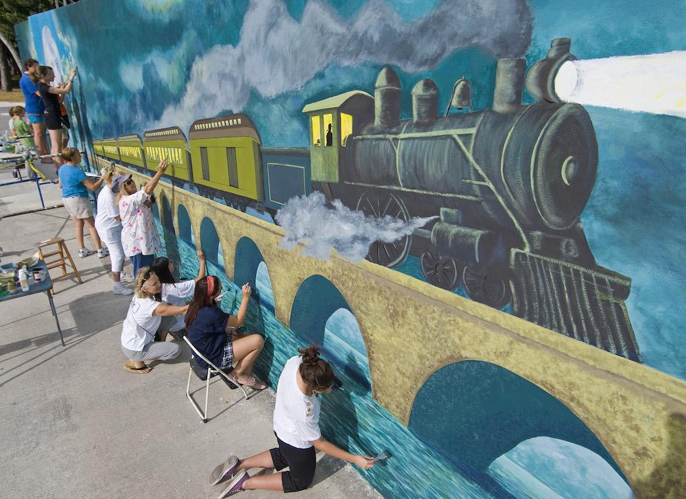 People painting a large mural of a steam train on a bridge.