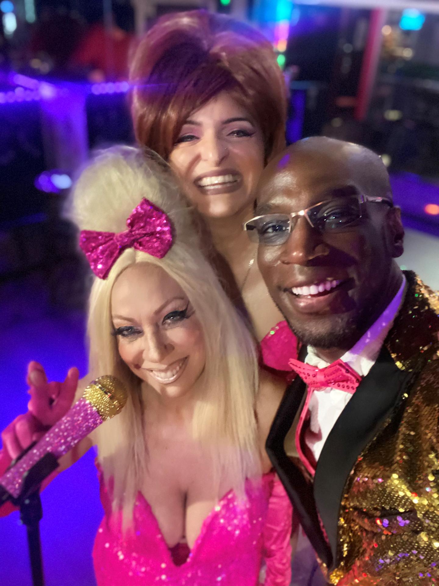 Three people smiling, dressed in glittery outfits, on a colorful stage.