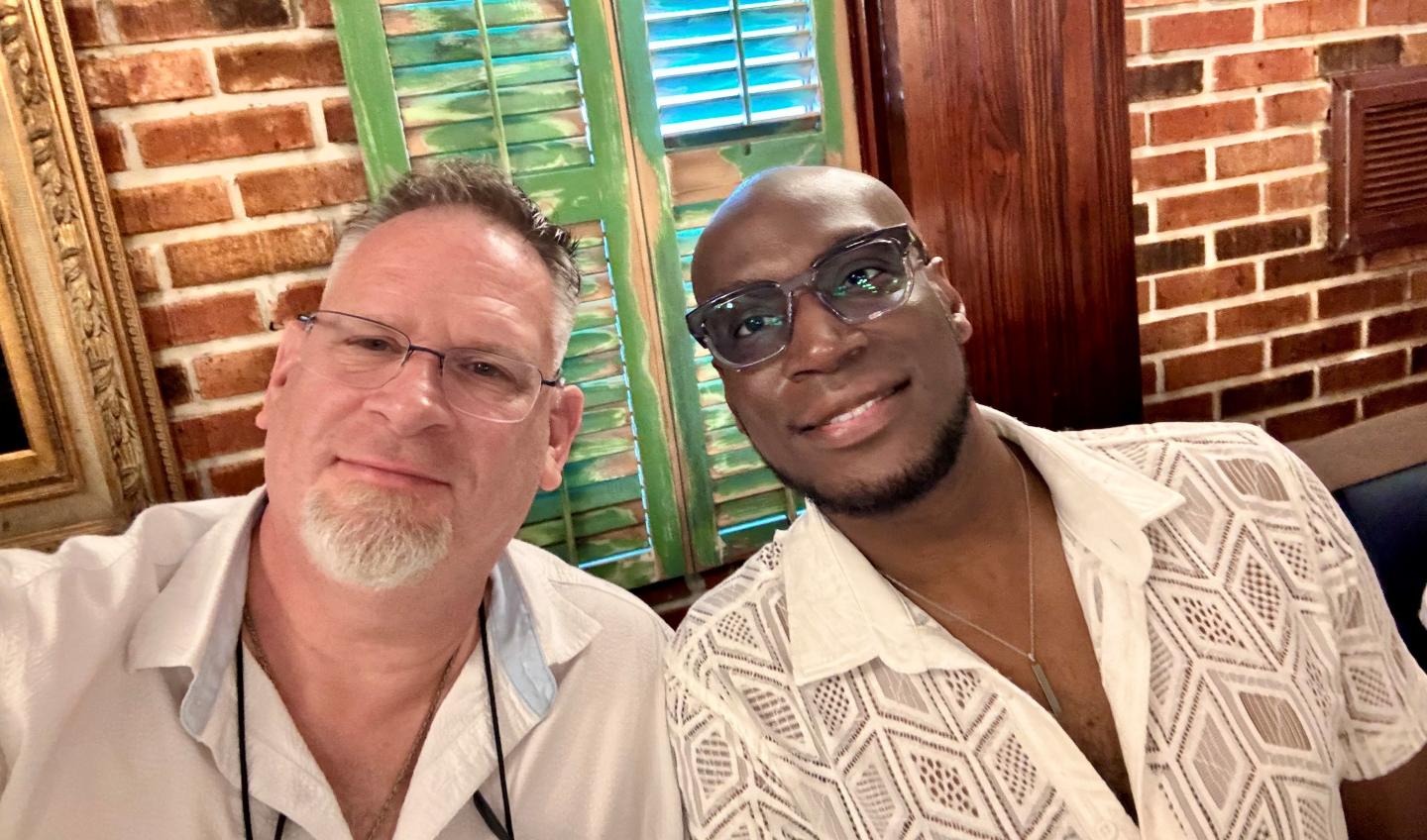 Two smiling men in a restaurant with brick walls and green shutters.