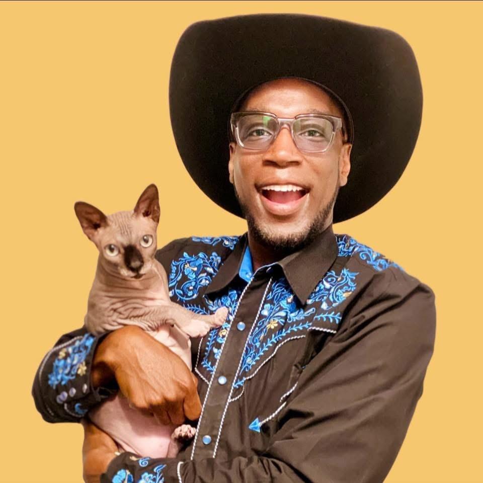 Cowboy hat man in blue shirt smiling, holding a hairless cat.