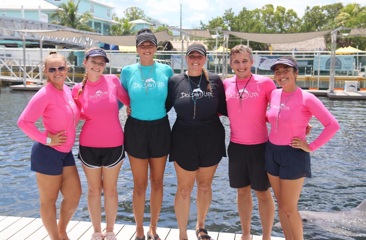 Six people in colorful shirts stand together smiling near a body of water.
