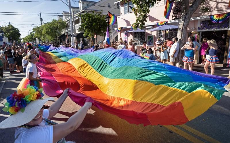 Crowd holding a large rainbow flag at a vibrant outdoor parade.
