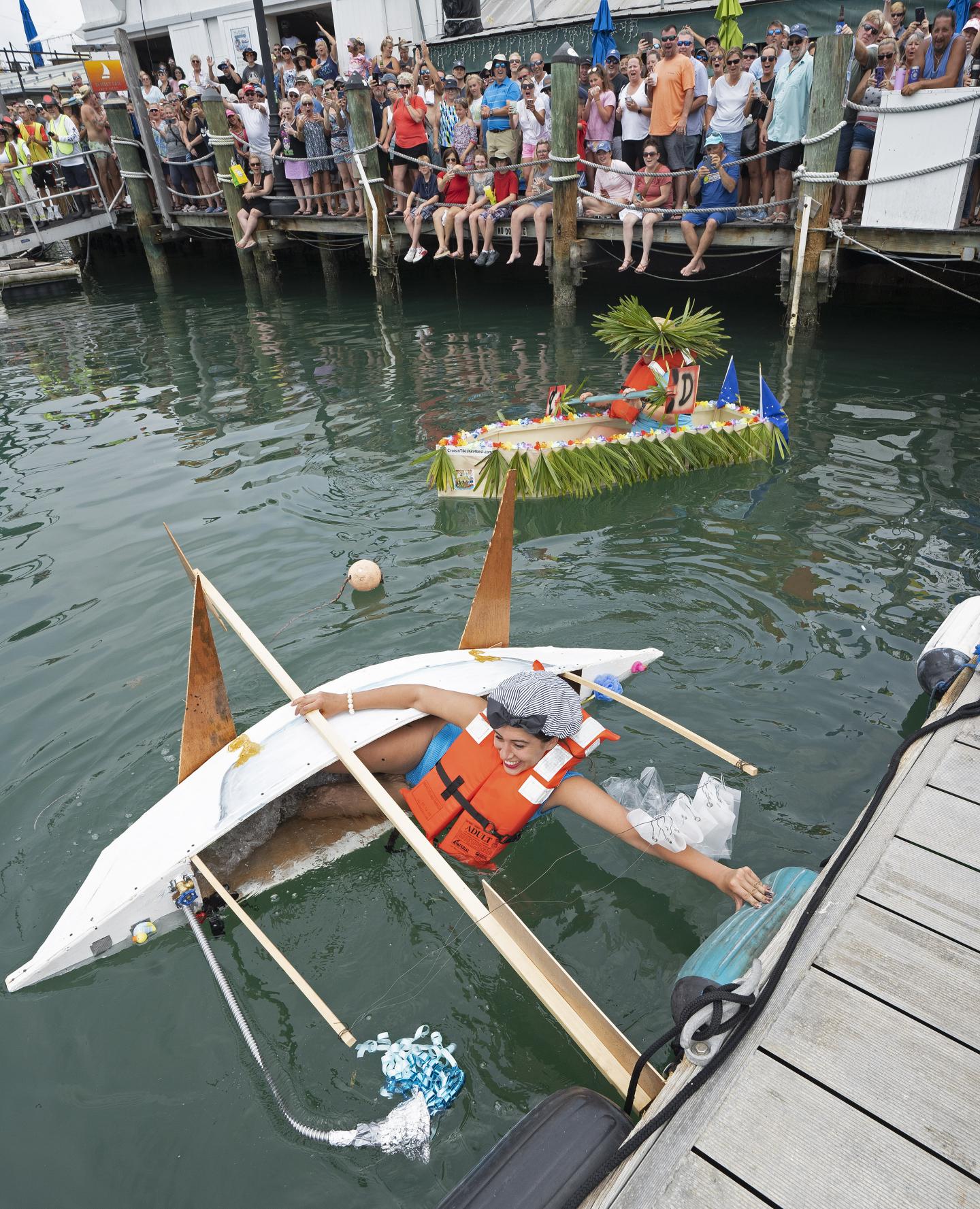 Man in a life vest in a sinking makeshift kayak, crowd watching from a dock.