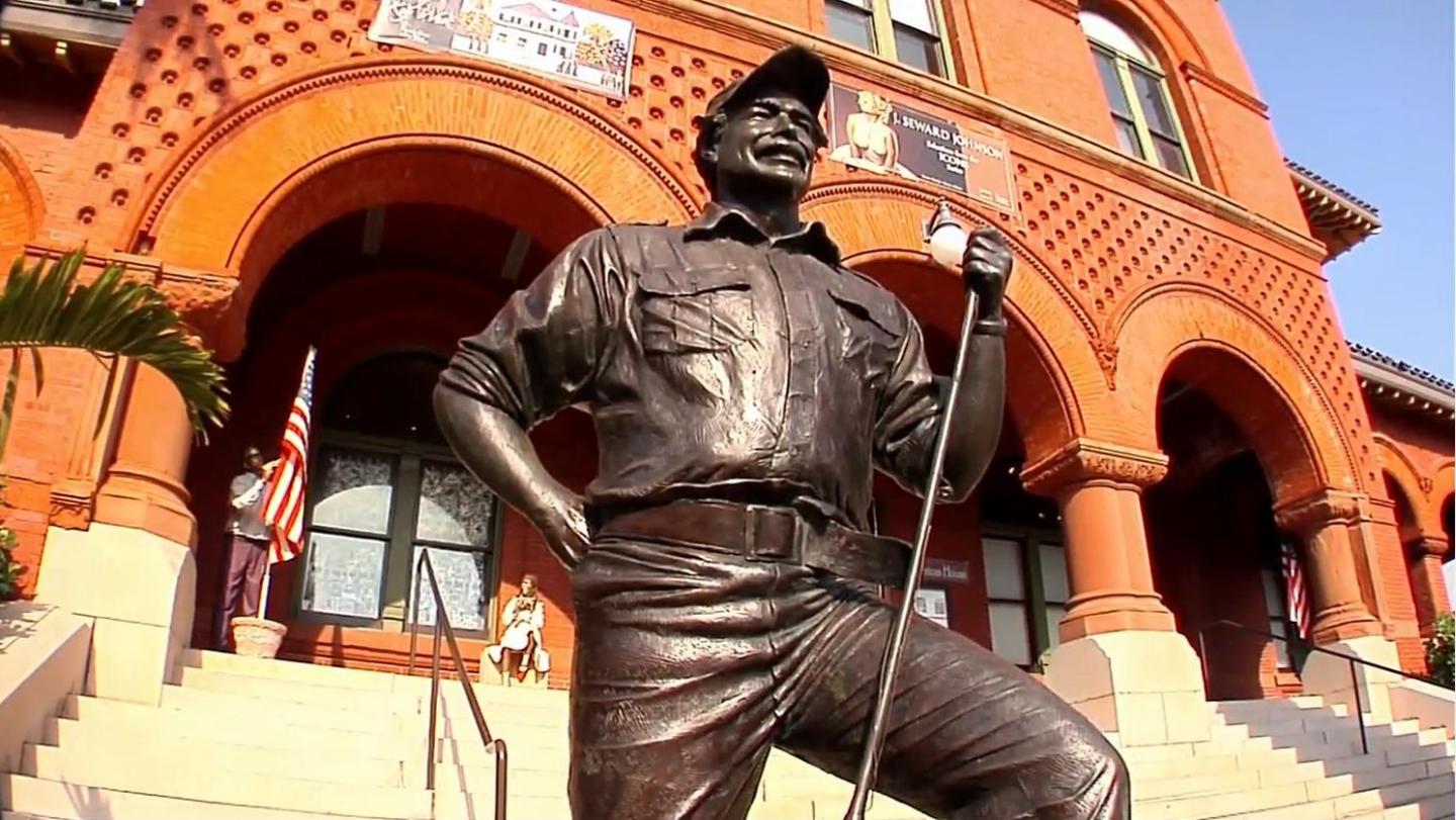 Bronze statue of a smiling man with fishing gear in front of a red brick building.