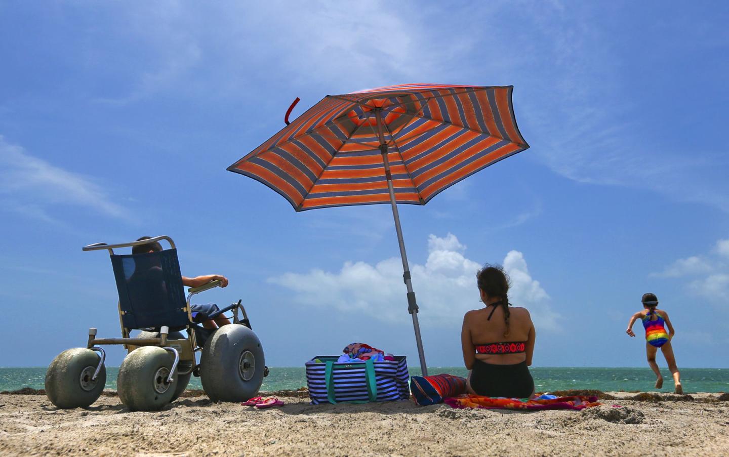 Beach scene with a wheelchair, striped umbrella, and two people enjoying the sun.