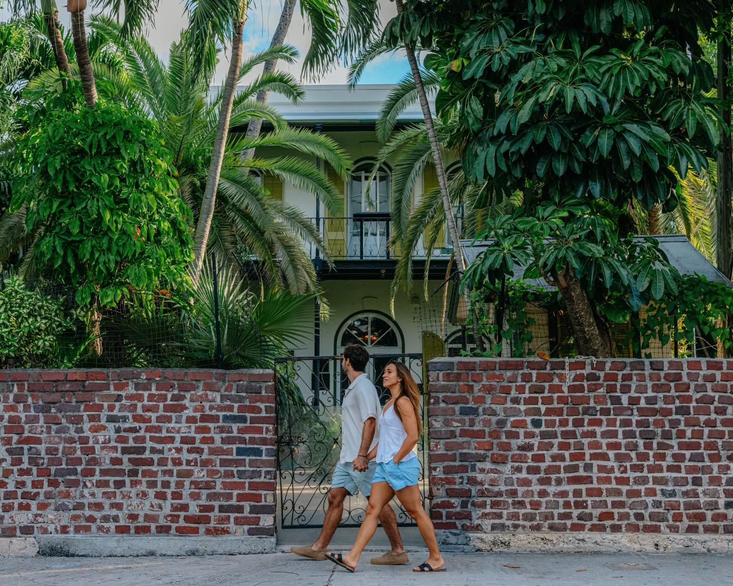 Couple walking past a brick wall with lush palm trees and a house behind.