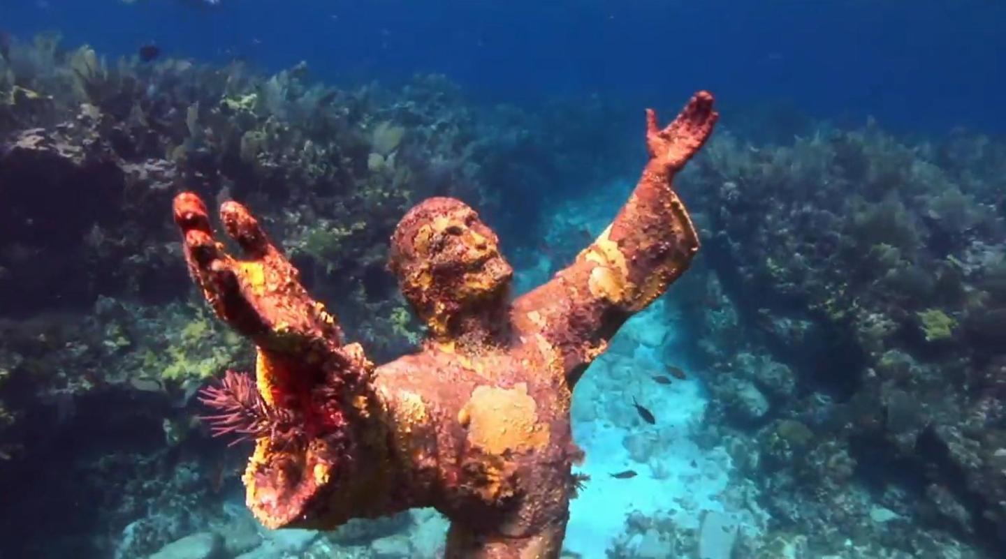 Christ of the Abyss underwater statue with outstretched arms, surrounded by coral and fish.