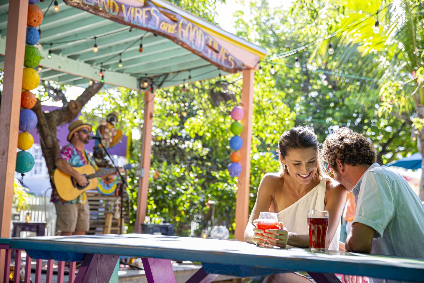 Couple sitting at a picnic table under colorful canopy, a musician plays guitar nearby.
