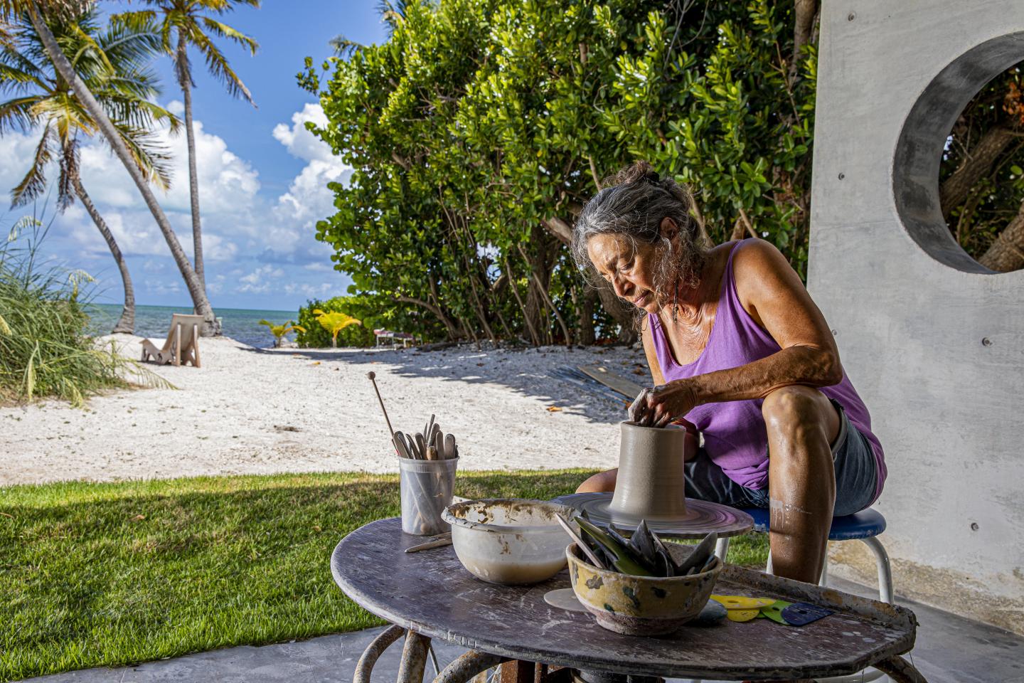 Woman sculpting clay pot, seated on outdoor patio near ocean and palm trees.