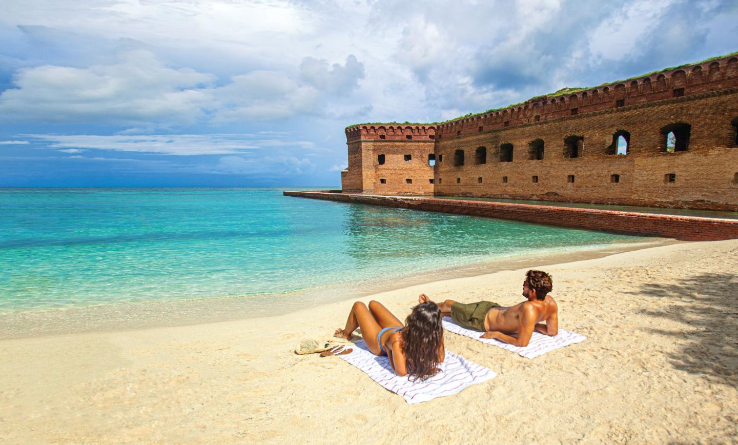 Two people sunbathe on a sandy beach near a historic brick fort by turquoise water.