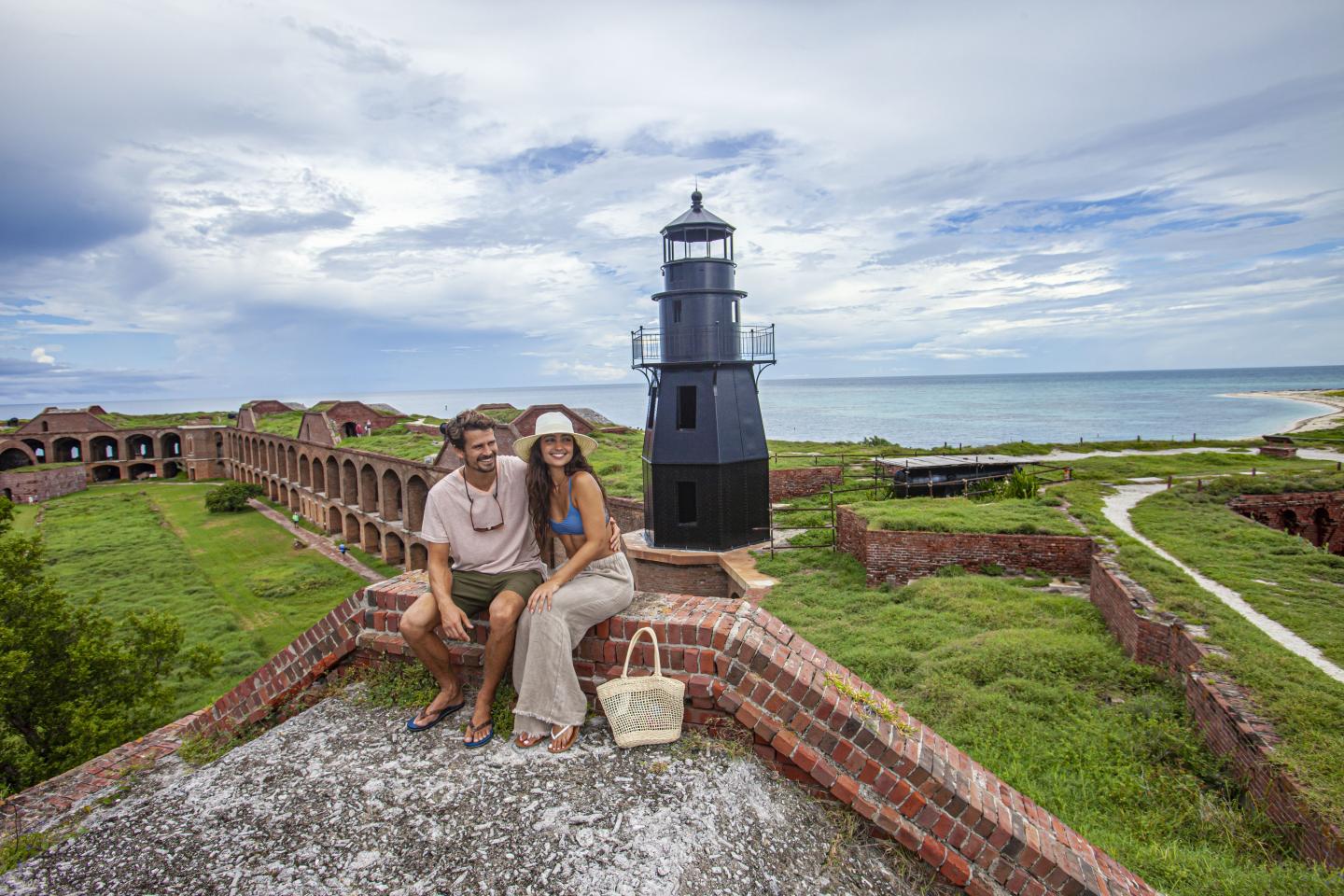 Couple sitting by lighthouse at Fort Jefferson, overlooking ocean and greenery.