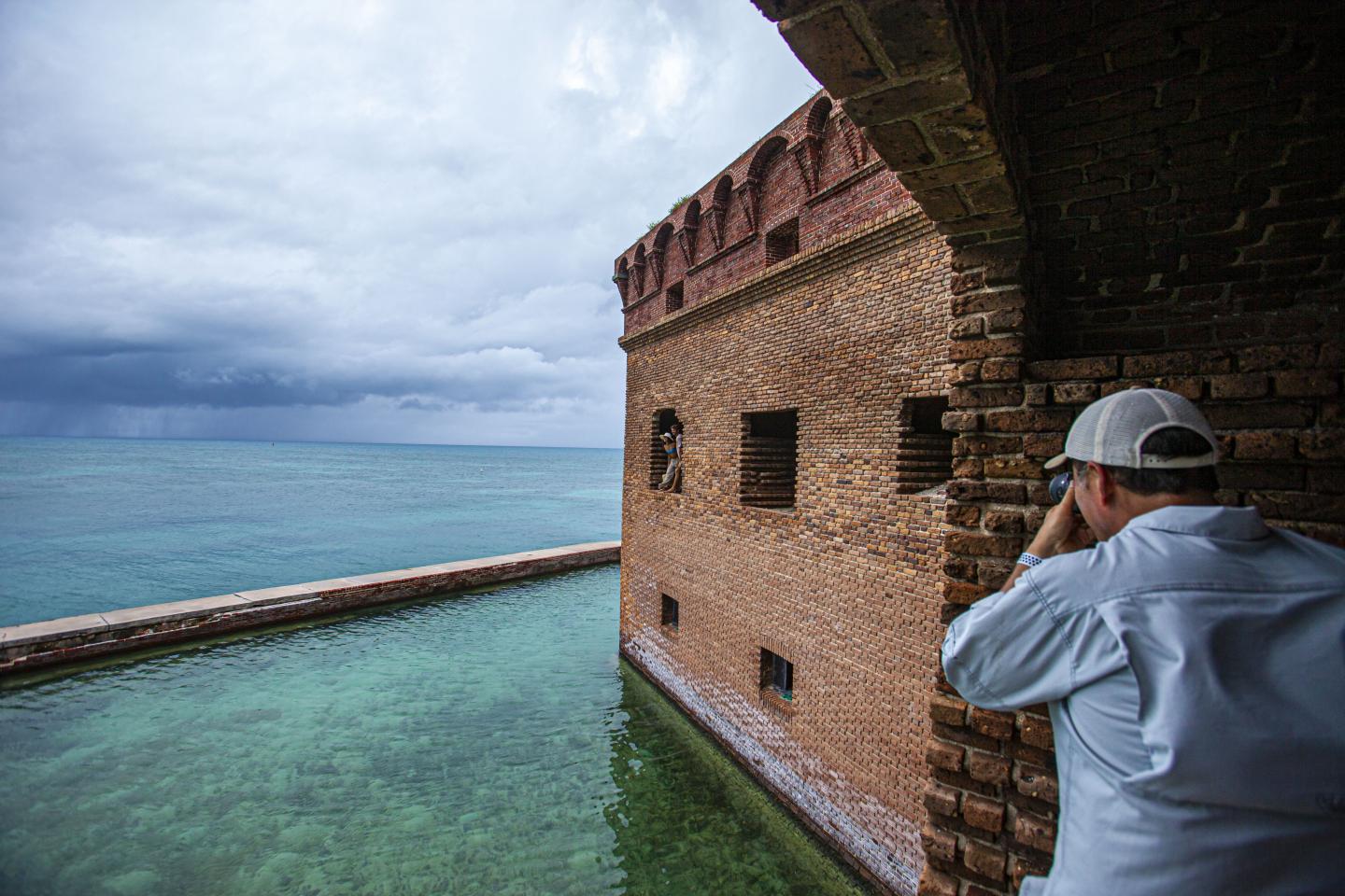 Fort wall by the sea with overcast sky, person photographing the view.
