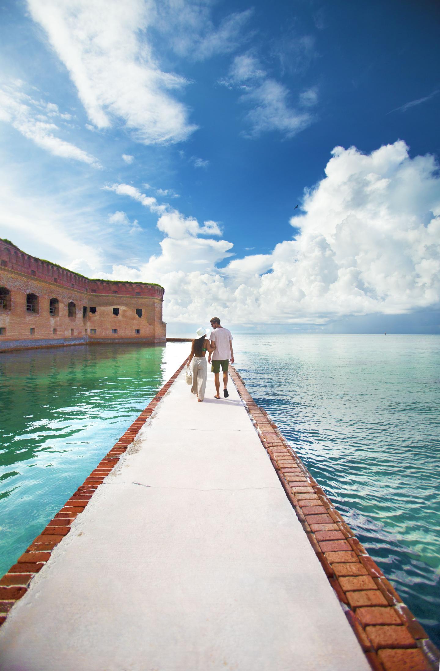 A couple walks on a pier toward a brick fort under a bright blue sky.