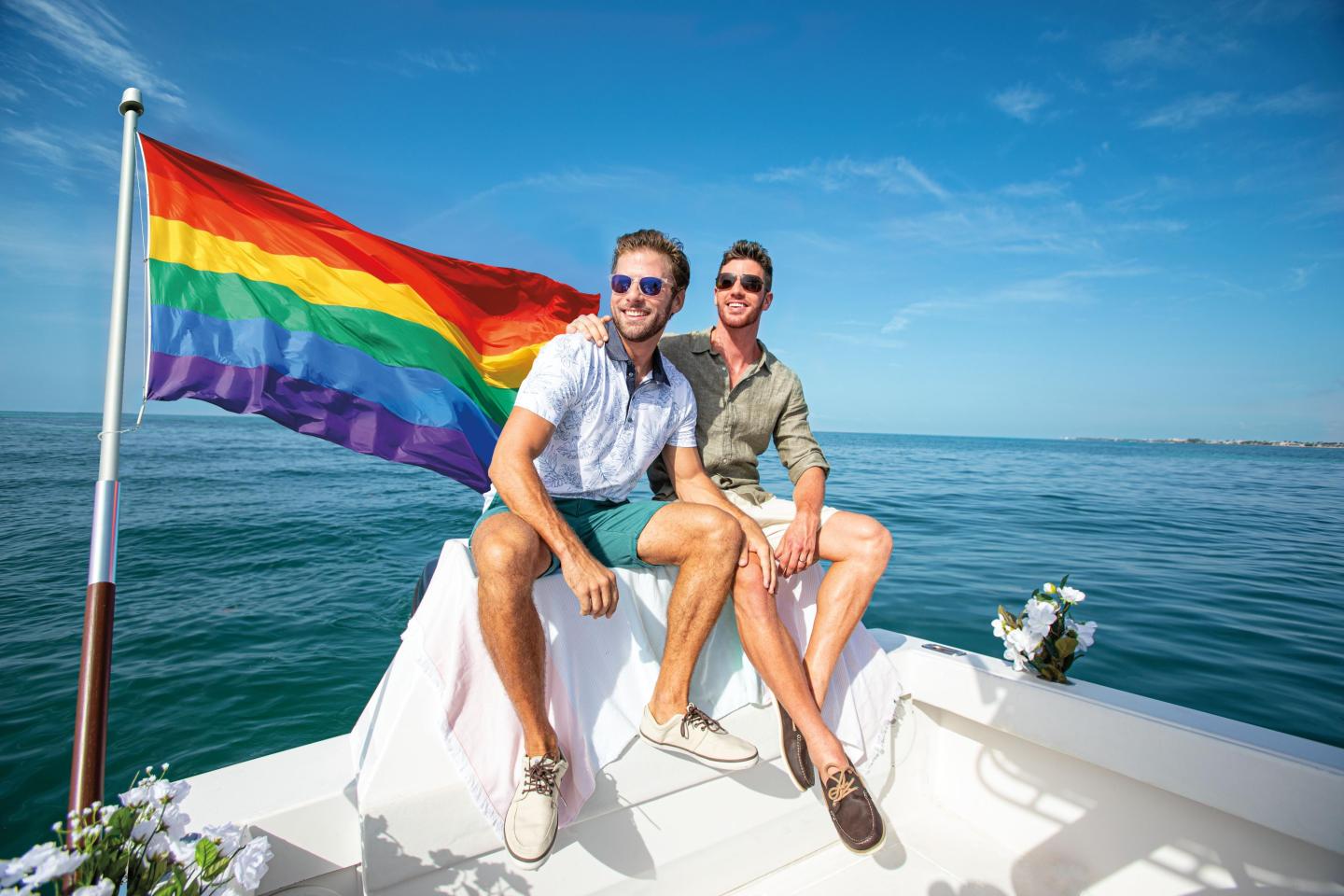 Two people smiling on a boat with a rainbow flag, under a clear blue sky.