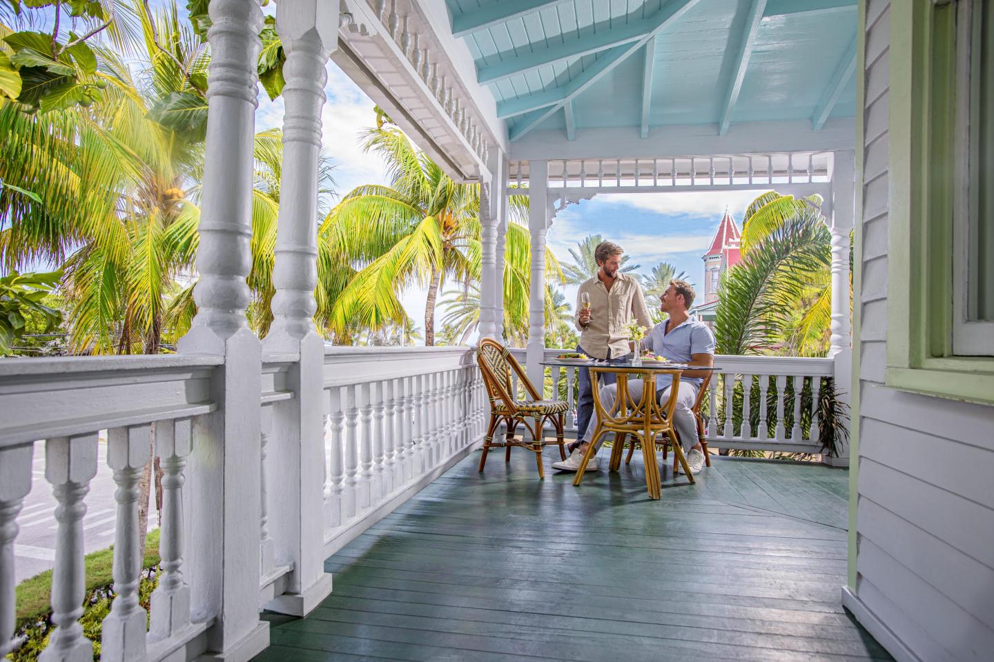 Veranda with two people at a table, surrounded by palm trees.
