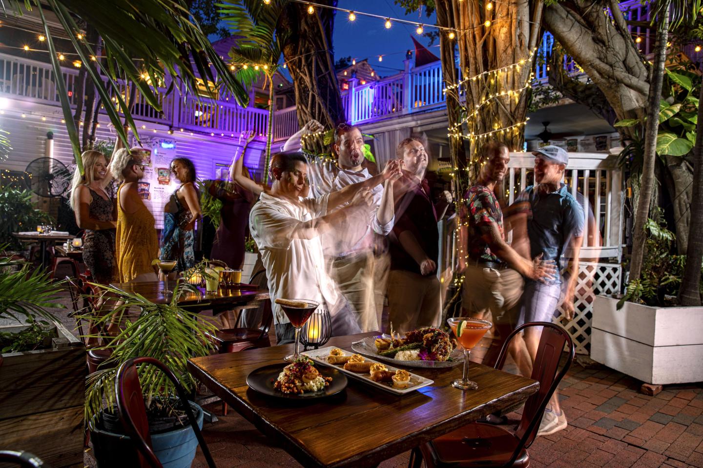 Outdoor party with people dancing under string lights, food on a table in foreground.